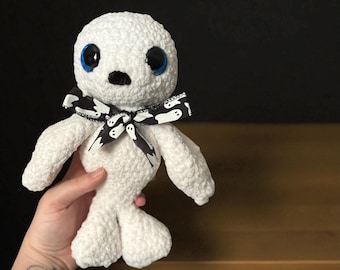 May include: A white crocheted ghost doll with large blue eyes and a black bow tie patterned with white ghosts. The doll has a rounded head, small black nose, and a body with arms and a tail-like bottom.