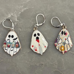 May include: Five silver-toned leverback earrings featuring ghost-shaped charms. The ghosts display various designs: one reading a book with a flower crown, one with iced coffee cups, one with red splatters, one making a peace sign, and one with flowers.