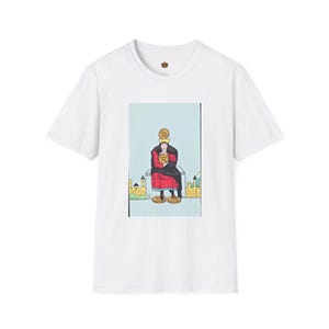 Puede incluir: Una camiseta blanca de manga corta con una ilustración gráfica centrada de una figura con corona y túnicas rojas y negras, sentada sobre un bloque de piedra sosteniendo una moneda de oro, con monedas adicionales bajo los pies y sobre la cabeza.