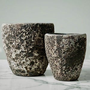 May include: Two planters with a rough, porous, and textured surface in shades of dark charcoal, brown, and off-white. The larger planter is wider and taller, while the smaller planter is narrower, both featuring a tapered, cylindrical shape.