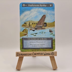 May include: Hand-painted artwork of a Zephyrranne Airship card. The card features a beige airplane with a brown tail, set against a blue sky with clouds. Text includes "Elite Mortals take to the skies" and "Airborne, Movement +2". The card is displayed on a small wooden easel.