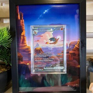 May include: Framed Charizard ex Pokémon card. The card features a red and orange dragon against a desert landscape background. The frame is black, and the artwork is protected by a clear cover. The card's text includes "Brave Wing" and "Explosive Vortex."