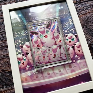Puede incluir: Una obra de arte enmarcada con una carta Pokémon Wigglytuff. La carta está dentro de una funda protectora y centrada sobre una impresión de múltiples personajes de Jigglypuff. El marco es blanco.