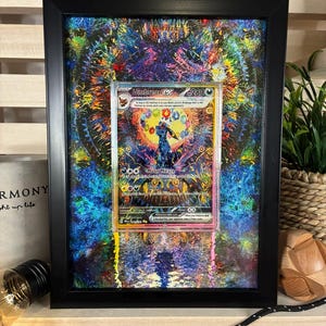 Op de afbeelding: Ingelijste Umbreon EX ruilkaart. De kaart zit in een doorzichtige plastic hoes en is geplaatst tegen een kleurrijke, abstracte achtergrond. Het zwarte frame past bij het ontwerp van de kaart. De kaart toont een donkerblauw wezen.