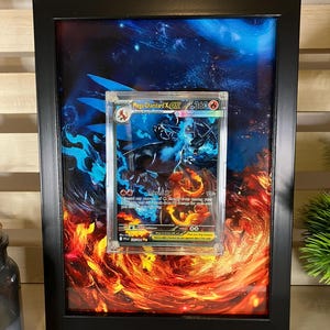 Könnte beinhalten: Gerahmte Mega Charizard EX Pokémon-Karte. Die Karte befindet sich in einer transparenten Hülle, vor einem feurigen Hintergrund aus orangefarbenen und roten Flammen. Der schwarze Rahmen umgibt das Kunstwerk.