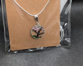 Puede incluir: Un collar plateado con un colgante redondo que presenta un árbol en miniatura con flores rosas. El colgante está encapsulado en resina transparente. El collar está empaquetado con una tarjeta marrón y una etiqueta verde que dice "JensGiftGarden".