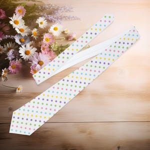 Pode incluir: Uma gravata branca com um padrão de bolinhas coloridas em amarelo, roxo, verde e rosa. A gravata está sobre uma superfície de madeira ao lado de um buquê de flores.