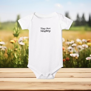 以下が含まれることがあります： 白い半袖ベビーボディスーツ、スナップ留め。前面には「Tiny But Mighty」の文字が黒でプリントされています。ボディスーツは、花と緑のぼやけた背景を持つ木製の表面に表示されています。