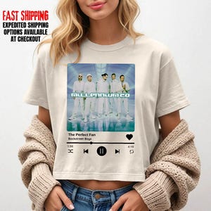 May include: Cream-colored crop top featuring a graphic of the Backstreet Boys' album "Millennium 2.0" with the song "The Perfect Fan." The design mimics a music player interface, with play/pause and skip controls. The top has short sleeves.