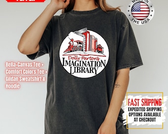 Może przedstawiać: Ciemnoszary t-shirt z krótkim rękawem z okrągłą grafiką przedstawiającą czerwony pociąg parowy wyłaniający się ze stosu książek. Napis głosi Dolly Parton's Imagination Library. Koszulka ma wyblakłe wykończenie w stylu vintage.
