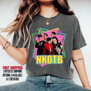 May include: Dark gray t-shirt featuring a colorful graphic of the band NKOTB. The graphic includes the band members in front of a pink brick wall with a neon green and blue frame. The band's name is in yellow and pink.