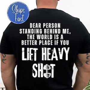 Könnte beinhalten: Schwarzes T-Shirt mit weißem Text: „LIEBE PERSON, DIE HINTER MIR STEHT, DIE WELT IST EIN BESSERER ORT, WENN DU SCHWER HEBST.“ Ein blauer Kreis mit „Schneller Versand“ befindet sich oben links. Das Shirt hat eine Hantel-Grafik.