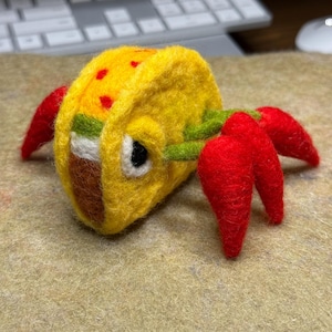 May include: A needle-felted sculpture shaped like a taco with a yellow shell featuring red spots, a brown filling, and a white layer. The taco has a large white and black eye and four bright red crab-like claws extending from the sides.