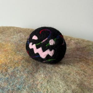 Puede incluir: Una esfera de lana negra fieltrada con aguja que presenta una boca de color rosa claro, dentada y cosida, junto a dos pequeños ojos circulares de color rosa claro. La superficie de la lana negra tiene hilos finos en tonos azul, verde y rojo.