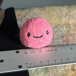 Könnte beinhalten: Ein kleines, rosa gefilztes Wesen mit einem lächelnden Gesicht. Der handgefertigte Artikel hat einen Durchmesser von etwa 4 cm. Die Augen und der Mund sind schwarz. Ein Metalllineal mit Zoll- und Zentimetermessungen befindet sich im Vordergrund.