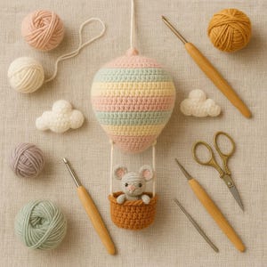 Puede incluir: Un globo aerost&aacute;tico de ganchillo hecho a mano en colores pastel con un peque&ntilde;o rat&oacute;n en una cesta. La imagen incluye ovillos de lana, ganchillos, tijeras y piezas de ganchillo en forma de nube. El globo est&aacute; rayado en rosa, amarillo y azul.