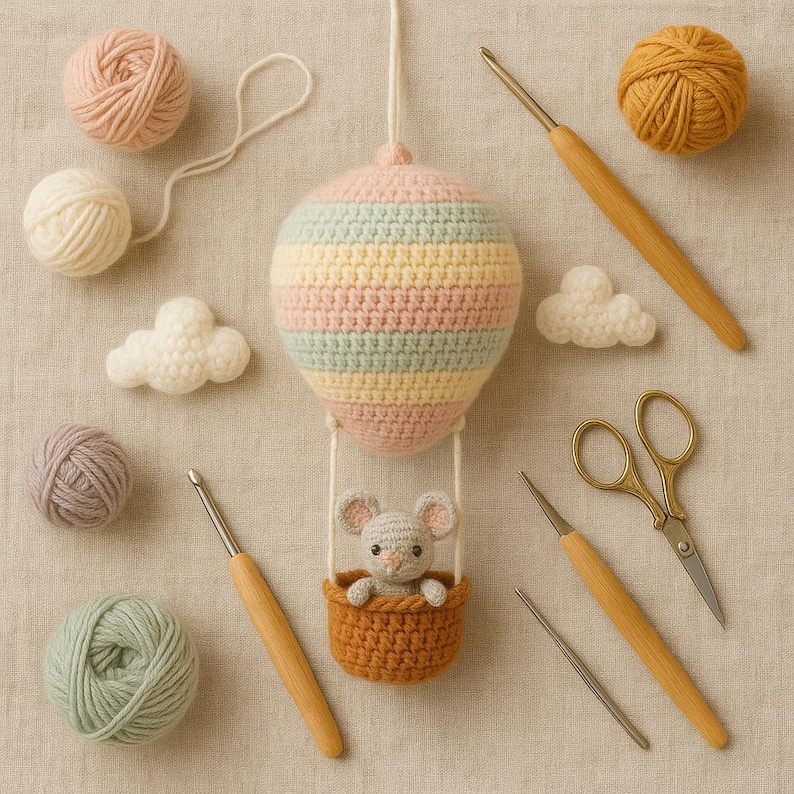 Puede incluir: Un globo aerost&aacute;tico de ganchillo hecho a mano en colores pastel con un peque&ntilde;o rat&oacute;n en una cesta. La imagen incluye ovillos de lana, ganchillos, tijeras y piezas de ganchillo en forma de nube. El globo est&aacute; rayado en rosa, amarillo y azul.