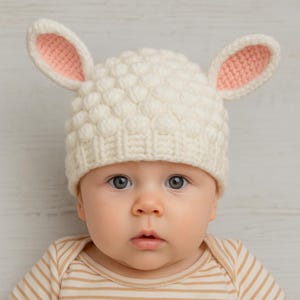 May include: A baby wearing a white knitted hat with pink inner ears, resembling a lamb. The hat has a textured, bobble-like pattern. The baby is also wearing a striped, tan and white onesie.