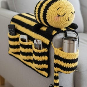 Pode incluir: Um organizador de sofá de crochê feito à mão em forma de abelha. O organizador listrado amarelo e preto tem vários bolsos para controles remotos, telefones e outros itens. A abelha tem um rosto sorridente e asas transparentes. Projetado para o braço do sofá.