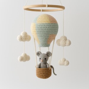Puede incluir: M&oacute;vil de beb&eacute; hecho a mano con ganchillo, con un globo aerost&aacute;tico en colores pastel, un rat&oacute;n gris en una cesta marr&oacute;n y nubes blancas y esponjosas. El m&oacute;vil cuelga de un aro de madera.