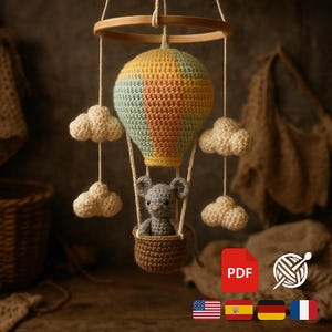 Könnte beinhalten: Handgefertigtes Häkelmobile mit einem Heißluftballon in Gelb, Pfirsich und Blau. Eine graue Maus sitzt in einem braunen Korb unter dem Ballon, umgeben von weißen Wolkenakzenten. Das Mobile hängt an einem Holzring.