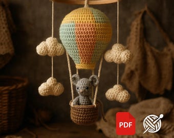 May include: A handmade crochet mobile featuring a hot air balloon with a yellow, peach, and blue design. A gray mouse sits in a brown basket suspended below the balloon, surrounded by white cloud accents. The mobile hangs from a wooden ring.