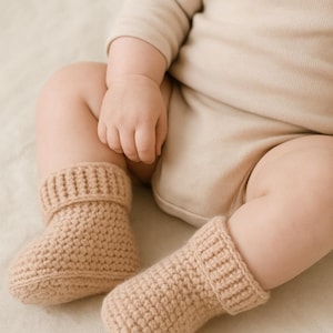May include: A pair of beige crochet baby booties with ribbed cuffs. The booties are made of a soft, textured yarn. These booties are designed to keep a baby's feet warm. Part of a baby clothing set.