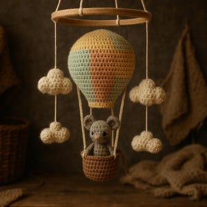 Puede incluir: M&oacute;vil de beb&eacute; hecho a mano con un globo aerost&aacute;tico de ganchillo amarillo, verde y marr&oacute;n. Un rat&oacute;n gris est&aacute; sentado en la cesta marr&oacute;n, y nubes color crema cuelgan debajo. El m&oacute;vil est&aacute; suspendido de un anillo de madera.