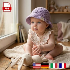 Puede incluir: Un bebé con un sombrero de cubo de ganchillo lavanda y un body con estampado floral está sentado en una manta beige. Un conejito de peluche está frente al bebé. Un icono PDF está en la esquina superior izquierda.