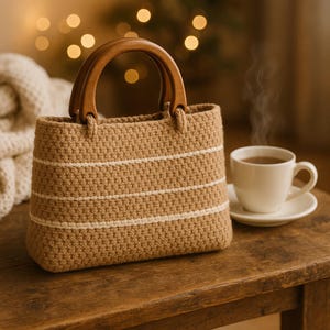 May include: A crocheted handbag with a wooden handle, featuring horizontal white stripes on a tan background. A white teacup and saucer sit nearby on a wooden surface. The bag has a rectangular shape.