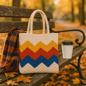 May include: A cream-coloured tote bag with a chevron pattern in yellow, red, and blue sits on a park bench. A plaid scarf and a coffee cup are also on the bench. The background is blurred with autumn foliage.