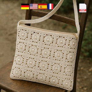 Puede incluir: Un bolso de crochet color crema con un patrón cuadrado y detalles de perlas. El bolso tiene cierre de cremallera y una correa larga. El bolso está sobre una silla de madera.