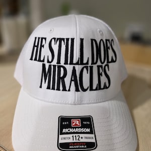 以下が含まれることがあります： 黒い文字で「HE STILL DOES MIRACLES」と書かれた白いRichardson 112+トラッカーハット。カーブしたつばとメッシュバックが特徴です。調整可能で、Richardson Flexfitのロゴが付いています。