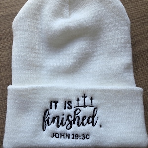 以下が含まれることがあります： 白いニット帽。黒い糸で「IT IS finished.」と「JOHN 19:30」が刺繍されています。柔らかく、質感のある素材でできており、つばが折り返されています。