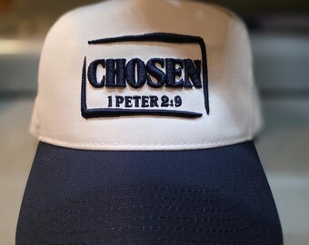 May include: A baseball cap with a white crown and a navy blue brim. The cap features the word "CHOSEN" embroidered in navy blue, along with the text "1 PETER 2:9" in a rectangular design.