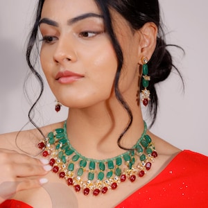 Könnte beinhalten: Ein grünes und rotes Perlenkette- und Ohrring-Set. Die Halskette hat smaragdgrüne Steine und rote Perlen mit goldfarbenen Akzenten. Die Ohrringe haben ein ähnliches Design mit grünen und roten Steinen.