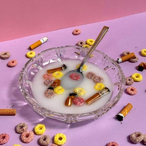 Pode incluir: Uma tigela de vidro transparente cheia de leite, cereais coloridos e pontas de cigarro. Uma colher de prata repousa no leite. O fundo é um gradiente de rosa e roxo.