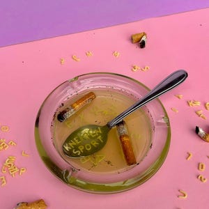 Könnte beinhalten: Ein klarer Glasaschenbecher mit zwei Zigarettenstummeln und einem Löffel darin. Auf dem Löffel steht "KNEIPEN SPORT". Der Aschenbecher steht auf einer rosa Oberfläche mit verstreuten Buchstaben aus Nudeln und Zigarettenstummeln.