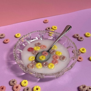 Op de afbeelding: Een heldere glazen kom gevuld met melk en kleurrijke ontbijtgranenringen. Een zilveren lepel rust in de melk, omringd door gele, roze en rode ontbijtgranen. De achtergrond is een verloop van roze en paars.