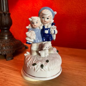 May include: A ceramic figurine featuring two children in blue and white outfits, standing on a white base with a small dog. The children are holding a doll and a basket. The figurine has a vintage aesthetic.