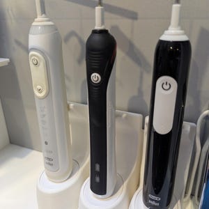 May include: Three electric toothbrushes in white, black, and black with red accents, each on a white charging base. The toothbrushes have power buttons and are labelled "Oral-B Braun".