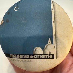 Peut inclure: Un objet rond en papier vintage avec un fond bleu et une illustration blanche d'une tour et de bâtiments. Le texte "maderas de oriente" est imprimé en noir, avec "MYRURGIA" en dessous.