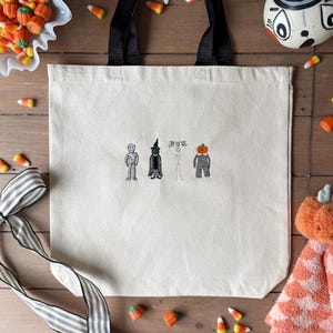May include: Cream-colored tote bag with black handles, featuring embroidered Halloween characters: a mummy, witch, skeleton, and pumpkin. Candy corn and a decorative pumpkin are scattered around the bag on a wooden surface.