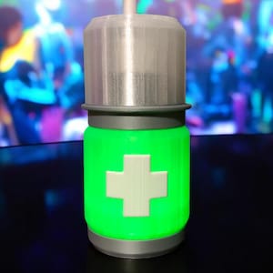 May include: A 3D-printed container with a silver cap and base. The body glows green and has a white cross symbol. The design suggests a medical or scientific theme. The container is cylindrical.
