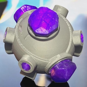 Könnte beinhalten: Ein graues, kugelförmiges Objekt mit sechs hervorstehenden Zylindern und vier großen, facettierten lila Kristallen. Das Objekt hat eine futuristische oder Science-Fiction-Ästhetik mit einem glatten, metallischen Aussehen.