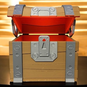 Könnte beinhalten: Eine hölzerne Schatzkiste mit offenem Deckel, die einen leuchtend roten Innenraum freilegt. Die Truhe hat Metallakzente, darunter Scharniere, Verschlüsse und eine Schlüssellochplatte. Die Außenseite ist hellbraun mit sichtbarer Holzmaserung.