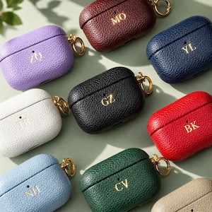 May include: A collection of textured leather earbud cases in various colors including purple, maroon, navy, white, black, red, light blue, dark green, and beige. Each case features a gold-toned metal ring and gold-embossed two-letter initials.