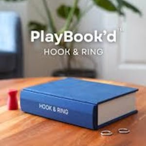 以下が含まれることがあります： 表紙にPlayBook'd HOOK & RING、背表紙にHOOK & RINGと書かれた青い本型の箱。小さな赤い木製のゲーム駒と2つの銀色の金属製リングが、青い長方形の箱の横の木製の表面に置かれています。