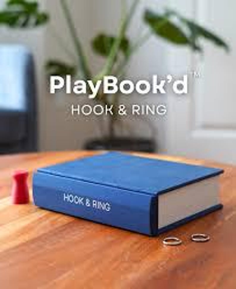 以下が含まれることがあります： 表紙にPlayBook'd HOOK & RING、背表紙にHOOK & RINGと書かれた青い本型の箱。小さな赤い木製のゲーム駒と2つの銀色の金属製リングが、青い長方形の箱の横の木製の表面に置かれています。