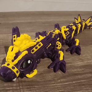 May include: A purple and yellow toy dragon with a detailed, mechanical design. The dragon has large eyes and a segmented body, with yellow accents on its head, back, and tail. The toy is made of plastic and sits on a wooden surface.